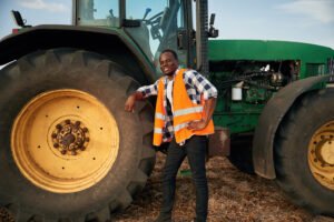 Leaning on the big wheel of tractor. Beautiful African American man is in the agricultural field.